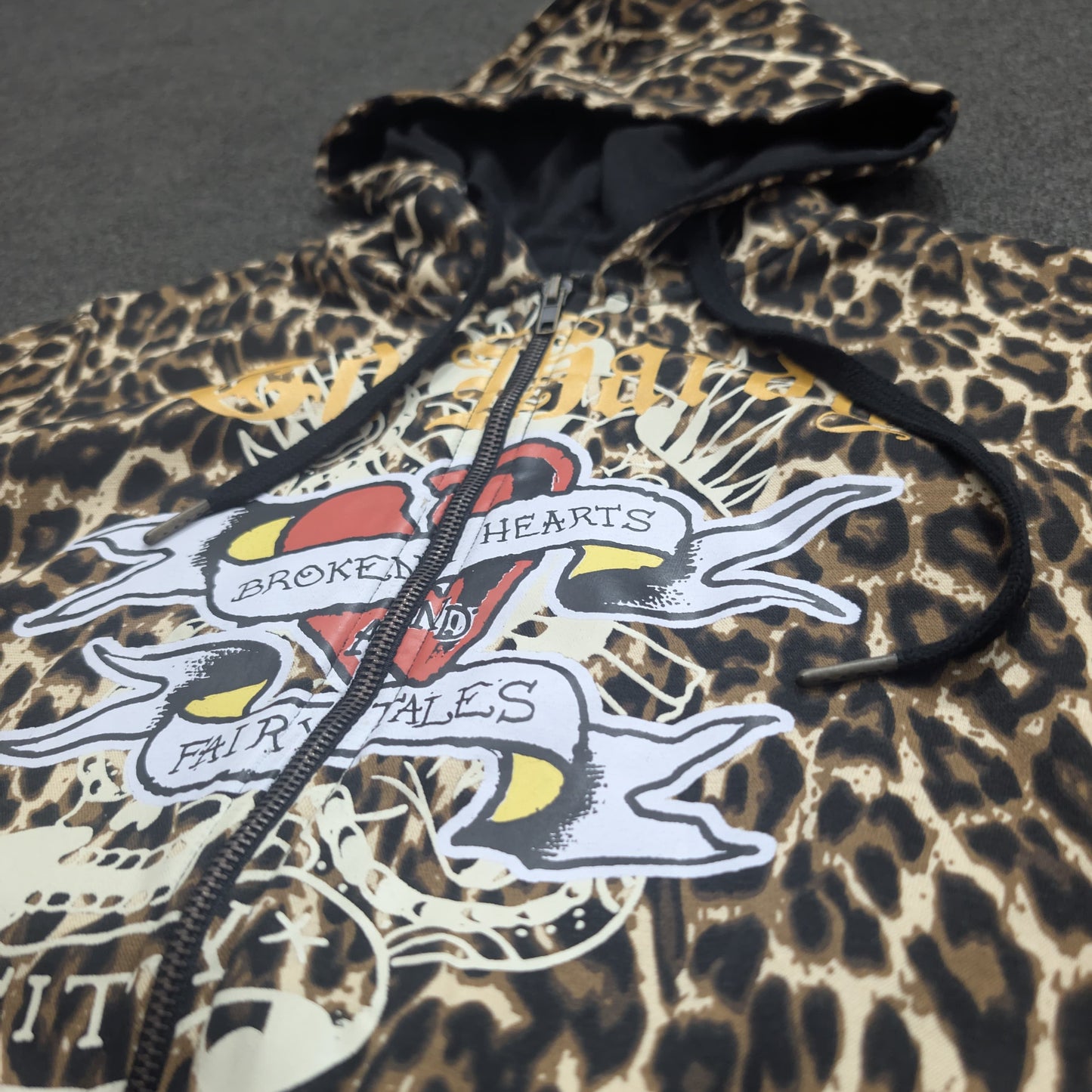 A leopard print zip-up hoodie featuring a large graphic print on the front with a broken heart, banners, and the words 'BROKEN HEARTS' and 'FAINT TALESS'.