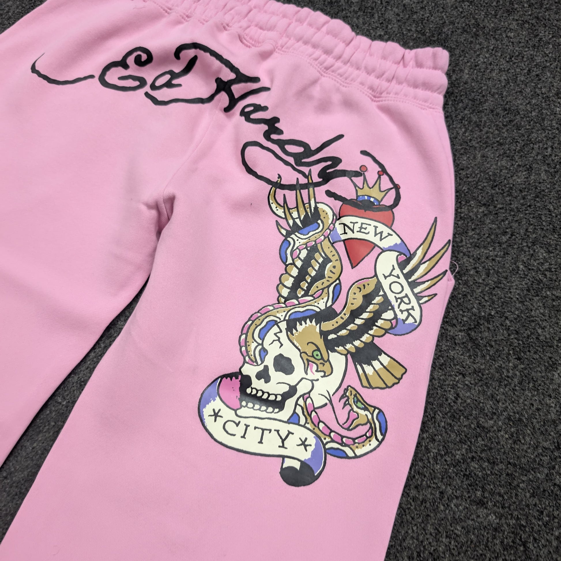 Pink sweatpants featuring a large Ed Hardy tattoo-style graphic on the leg, including a skull, eagle, and banner reading 'New York City', laid flat on a dark gray textured surface.